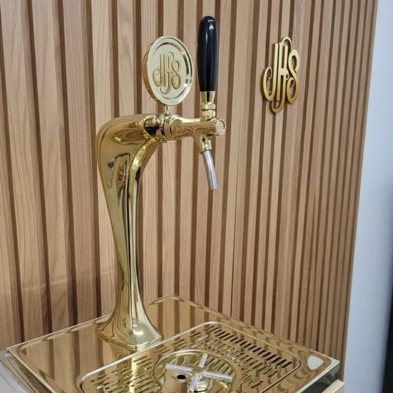 Chopeira dourada de uma torneira com o medalhão JHS, na parede de ripas de madeira com o monograma JHS